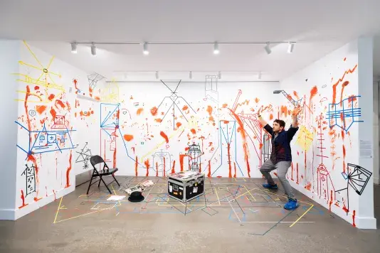 A man wearing a tie stands in a theatrical pose in a gallery: knees slightly bent, arms above his head, palms upturned. The walls and floor around him are marked with dense lines of colored tape and splotches and smears of red paint. A few props, like a hat, a typewriter, and a phone, sit on the floor too.