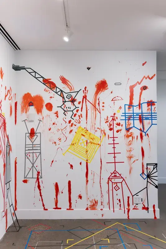 A white wall—densely marked with handprints and vertical trails of red paint, as well as obscure geometric figures made of colored tape—occupies most of the frame. The portion of the gray floor that is visible is also marked with boxes and brackets of multicolored tape.