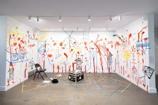 Three walls and the floor of a gallery are densely marked—with handprints and vertical smears of red paint, and with geometric figures and boxes made of colored tape. A few objects, including a metal folding chair, a shipping chest, a typewriter, and a phone, sit on the floor atop the tape markings.