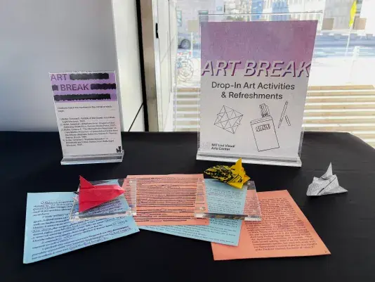 A table with a black tablecloth in front of a sunny window holds two clear acrylic signs with text that reads, "ART BREAK Drop-In Art Activities &amp; Refreshments." On the table, several pieces of paper with printed text and origami creations, including red, yellow, and white rocket ships, are arranged.