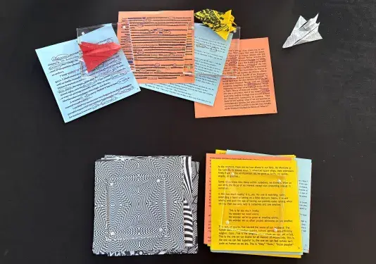 Various pieces of paper are displayed on a black surface. These include a blue sticky notes, a red paper rocket ship, an orange sticky note with black text, and a yellow sticky note topped with a small yellow paper sculpture and a white paper rocket ship. A black and white patterned sheet of paper and another yellow paper with text are also visible.