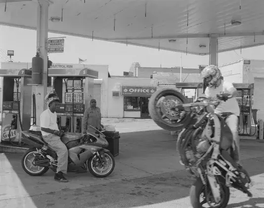 Black-and-white photo of a gas station where one motorcyclist performs a wheelie in the foreground while another sits on a sport bike near the pumps, with a third person standing nearby and the station office in the background.