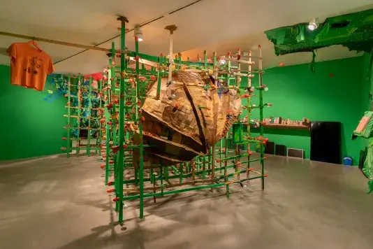 A large, textured, brown cardboard sculpture, is encased within a green wooden grid structure. The installation is set in a room with bright green walls, and an orange t-shirt hangs from the ceiling on the left side of the sculpture.