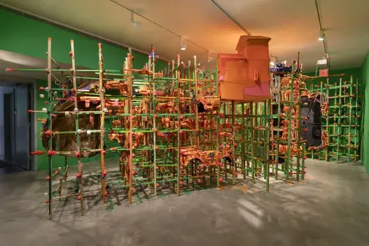 A large, intricate art installation fills a room with bright green walls. It features a dense, interconnected framework of vertical green poles and horizontal orange sticks. Integrated within this grid are various abstract, orange-toned structures resembling organic forms, toy guns, or small buildings and cars.