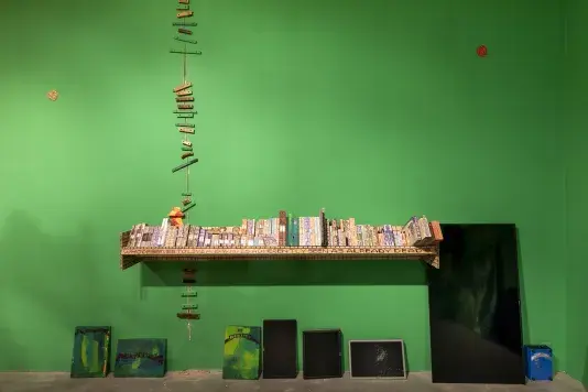 A bright green wall displays a long shelf filled with small, colorful, rectangular wooden objects, some with visible text. Below the shelf, three green paintings with text, four digital screens, and a blue container are arranged against the wall. To the left, a vertical line of small, colorful wooden blocks extends upwards connecting a seam in the wall.