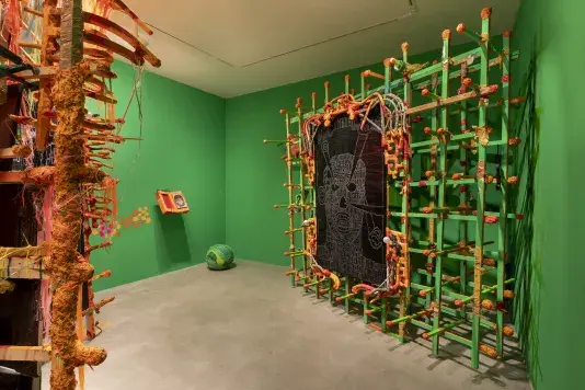 A room with bright green walls and a concrete floor. On the left, a tall, irregular structure of orange and brown textured materials stands. On the right, a large rectangular grid of green and orange rods frames a digital screen with white line drawings.