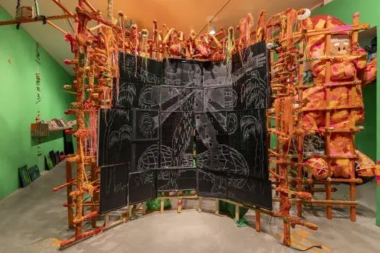 A large art installation features a structure made of wooden slats wrapped with orange tape and yarn in the center of the room. A gridded wall of digital screens with white chalk-like drawings are integrated into the structure, surrounded by various objects including an orange figure in the form of a Santa Claus inflatable.