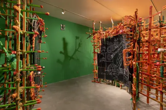 A large art installation features two complex, tunnel-like structures made of wooden slats wrapped with orange and green tape and yarn. A gridded wall of digital screens with white chalk-like drawings are integrated into the right structure, and a shadow of a structure is cast on the bright green wall behind.