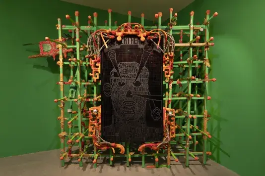 A large, rectangular sculpture features a digital screen at the center displaying white pixelated text in the shape of a face. This panel is encased in a green grid frame adorned with numerous colorful, wrapped elements in orange, red, and yellow, set against a bright green wall.