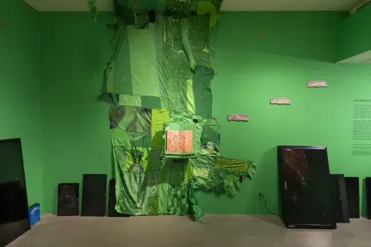 A large, mixed-media art installation covers a bright green wall, composed of draped and layered green fabrics with embedded reflective round objects and the form of an arm emerging from the ceiling. Two wooden blocks hinged together to form a book displays a reddish-orange, textured drawn surface. Three smaller, white, textured rectangles are mounted on the wall to the right and seven digital screens in various sizes lean against the wall on the floor.