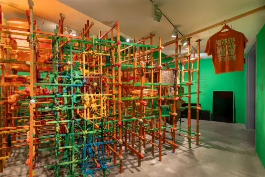 A large art installation features a dense, multi-level structure built from interconnected orange and green wooden sticks in a green walled room. Various colorful objects are woven into the framework. An orange t-shirt hanging from the ceiling displays the text: 'PAY FOR SOUP BUILD A FORT SET THAT ON FIRE'.