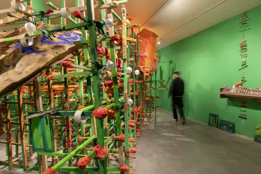 A large, complex sculpture made of green and orange painted wooden framework and various attached objects fills the left side of a gallery. Red, crumpled forms, white spheres, and a large brown textured element are visible in the structure. A blurred person wearing all black walks past a bright green wall. Books made of wooden blocks are lined in a row on a wooden shelf on the right side.