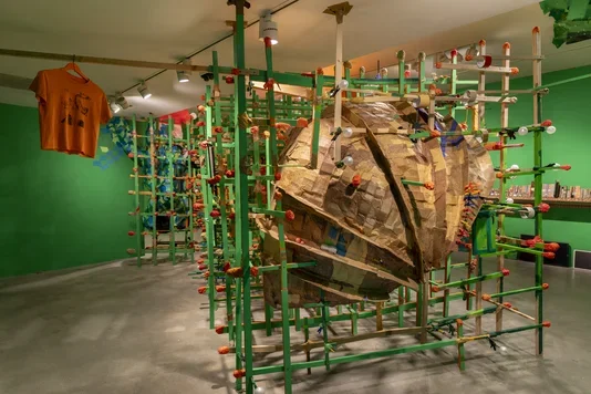 A large, textured, brown cardboard sculpture, is encased within a green wooden grid structure. The installation is set in a room with bright green walls, and an orange t-shirt hangs from the ceiling on the left side of the sculpture.