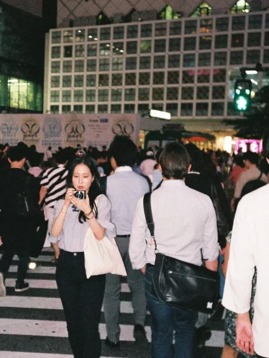 A woman holding a white tote bag is walking in a crowded crosswalk at night while examining a small camera.