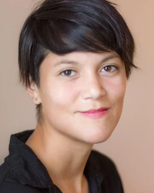 Headshot of a Chinese American woman with short dark hair and brown eyes wearing a black collared shirt in front of a beige wall.