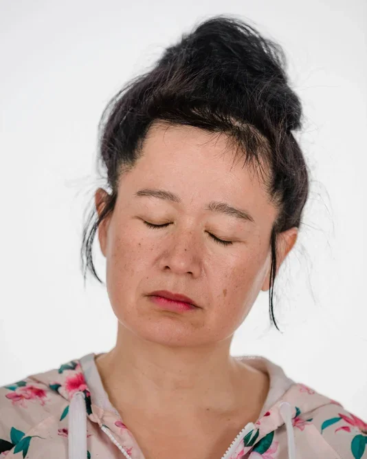Hito Steyerl pictured with a calm expression and closed eyes, has dark hair piled on her head and a floral zip up jacket.