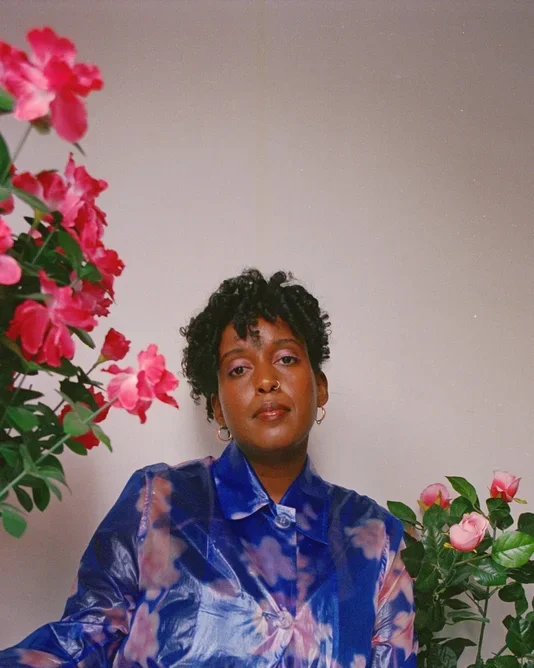 A woman with short dark curls and brown skin is wearing a blue printed silk blouse standing in between two colorful floral plants.