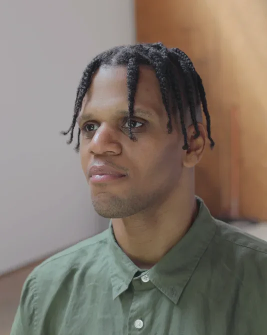 A man with brown skin and short dreads that fall over their forehead is sitting looking to the left of the frame wearing a green buttoned down shirt.