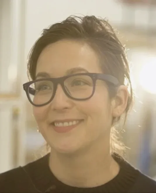 Headshot of a woman wearing a black shirt and black glasses smiling and looking passed the left side of the frame with sun glowing behind.