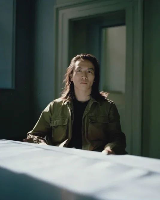 An asian man with long hair wearing a green military jacket and a black shirt is standing on a building balcony as the sun shines down.