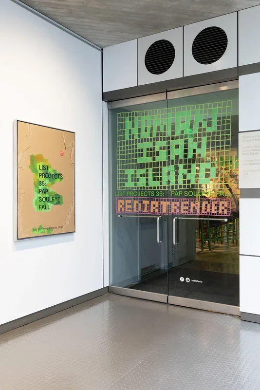 An entrance to a gallery features a cardboard sign on the left wall with green paint surrounding text reading 'LIST PROJECTS 35: PAP SOULEYE FALL' and dates. The glass door on the right has green grid-like text that reads ‘NO MAN IS AN ISLAND’ with orange text below reading ‘REDIRTRENDER’ above the door handles.