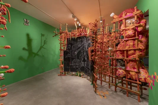 An art installation in a gallery with green walls and a gray floor. On the left, a vertical structure of textured orange organic shapes casts shadows. In the center, a curved black structure made of screens with white line drawings is supported by an orange stick framework, with a large, soft, orange and pink sculptural Santa with a cartoon-like face nestled within a dense orange stick structure to the right.