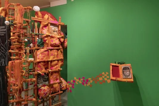 A large, colorful sculpture made of wood, fabric, and yarn stands on the left against a green wall. It is predominantly orange and pink, with complex woven and layered elements. To its right, an open, orange wooden book is mounted on the green wall, connected by a trail of small orange and pink rectangular paint gestures on the wall.