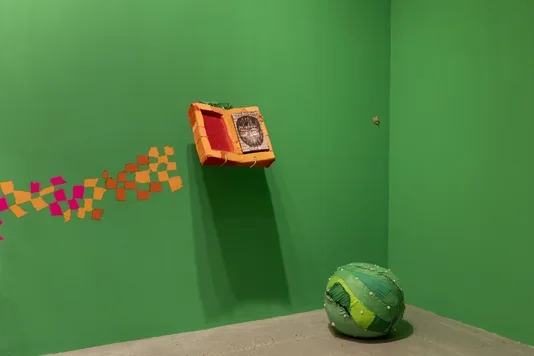 A bright green wall features a burst of painted orange and pink squares and an open orange fabric-covered box revealing a wooden book with a drawing of a figure’s head. On the concrete floor, a large green fabric sphere with white beads rests.