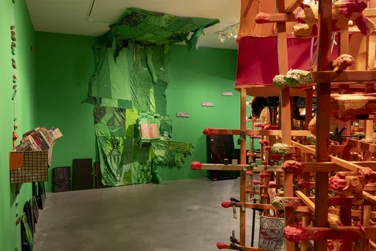 A gallery room with bright green walls and a concrete floor displays two large art installations. On the left, a large green fabric piece drapes from the ceiling, embedded with various objects. On the right, a tall, intricate wooden structure holds numerous orange, organic-shaped elements.