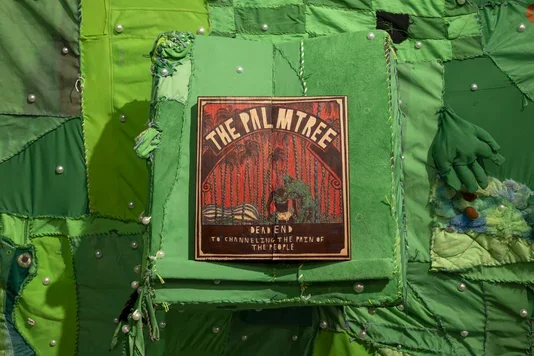 A dark wooden book, titled 'THE PALMTREE' and depicting palm tree silhouettes against a reddish-orange sky with a central figure, is centered on a square green fabric panel. The book also reads 'DEAD END TO CHANNELING THE PAIN OF THE PEOPLE. ' Both the panel and the surrounding patchwork of green fabrics are embellished with white pearl-like beads.