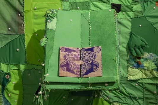 A large textile artwork made of various shades of green fabric patches sewn together, with white pearls embedded throughout. In the center, an open book with a light brown cover and purple and blue patterned pages rests on a raised green fabric. A green glove and other textured fabric elements are sewn onto the right side.
