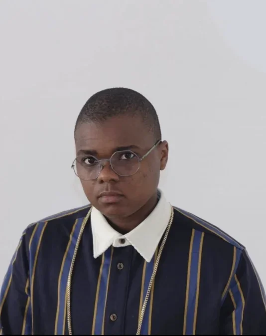 A person with brown skin and a buzz cut wearing a collared striped long sleeve shirt and glasses sits in front of a white wall looking at the camera.
