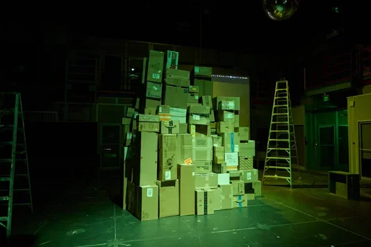 A large, tall pile of cardboard boxes stands in the center of a dark, green-lit room. A tall ladder is on the right, and a disco ball hangs from the ceiling.