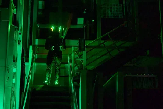 A person in dark clothing stands on a metal staircase with a large baseball bat in hand. Green lights glow from their head and feet, illuminating the dark, industrial setting.
