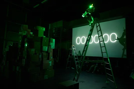 A dark room is illuminated by green light. A person on a tall ladder aims a green laser at a large screen displaying "00:00:00". To the left, a tall stack of cardboard boxes is visible, and several people sit on the floor in front of the screen.