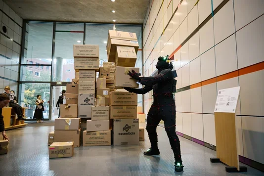 A figure in a dark, textured suit with glowing green accents on the head and feet removes a cardboard box to a tall, precarious stack in a brightly lit museum atrium.