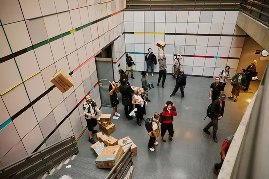 A group of people interact with cardboard boxes in a multi-level modern building. Some boxes are stacked on stairs, others are held, and one is in mid-air. The white tiled walls are decorated with horizontal colored stripes.