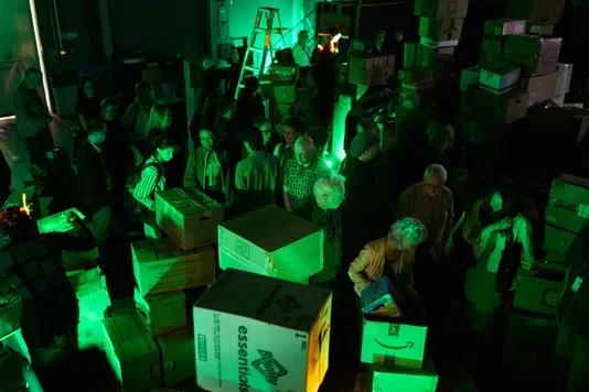 A large group of people stands amidst numerous cardboard boxes in a dimly lit room, illuminated by a strong green light. Boxes of various sizes are stacked and scattered throughout the space.
