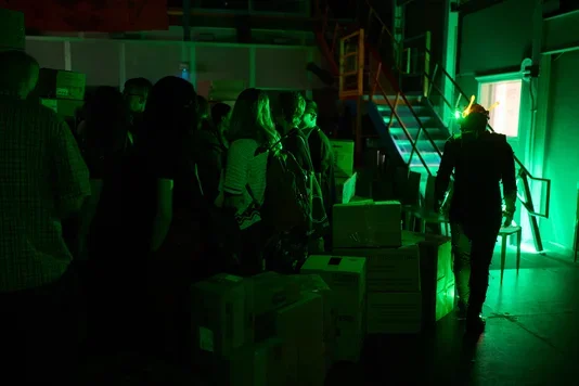 A person in dark attire with a glowing yellow headpiece walks away from a group of people in a dimly lit, green-illuminated industrial setting. Stacked boxes are in the foreground, and a metal staircase is visible in the background.
