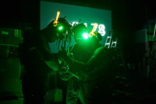 Three figures in dark, motion-capture-like suits with glowing green headgear stand facing each other in a dimly lit space. Small pearl-like dots are visible on their bodies, and the number 78 is projected in green light on a screen behind them.