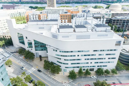Aerial view of a white building with lots of windows on the corner of two streets. In the background, there are a lot more buildings and the Charles river at the top of the frame.
