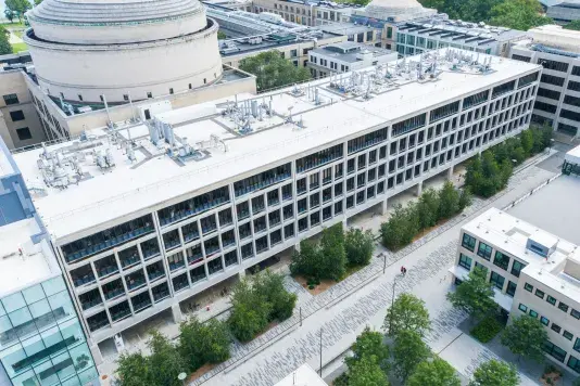 Aerial shot of a grey, white building with lots of rectangular windows situated on a block between lots of other interconnected buildings.