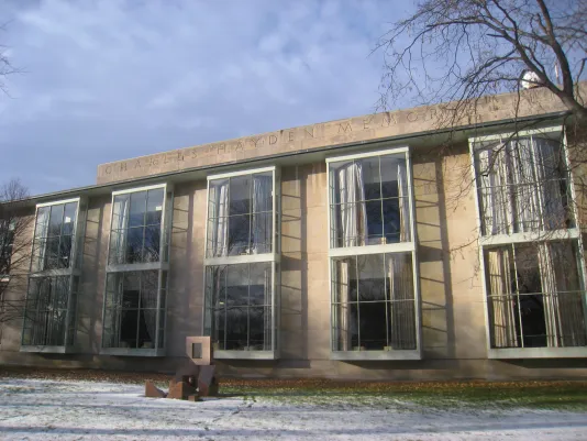 Grey building with 5 large vertical windows and a sculpture on the front lawn.