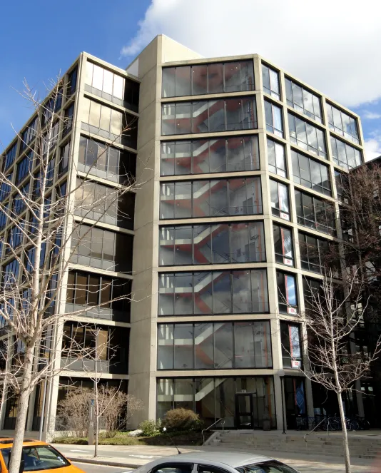 Vertical image of a grey building with lots of windows featuring a stairwell inside the center panel of windows.