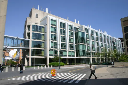 Building featuring lots of windows and a glass catwalk on the left. In the foreground is a street with a crosswalk and pedestrians.