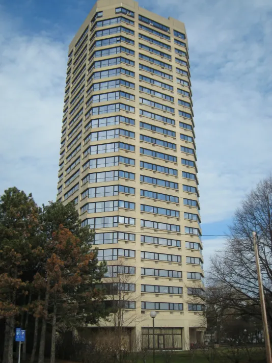Tall grey building with lots of floors and windows with a few trees in the left corner of the frame.
