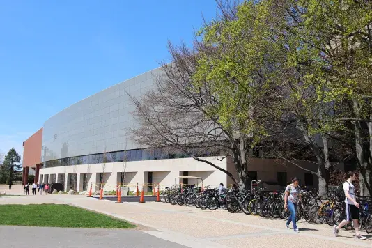 Grey and brick building with a horizontal floor of windows featuring a bike rack on the right with lots of bikes and people walking on the sidewalk.