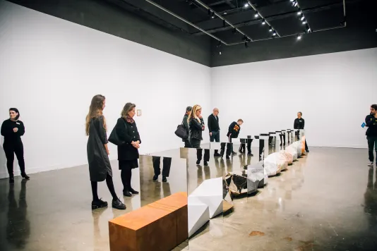 Six people stand scattered around a sculpture on the floor. The sculpture consists of a row of large rocks and geometric figures situated between mirrors.