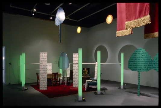 Green pillars surround a dining table and walls of text. Cut-out trees covered in text sit outside of the installation. 