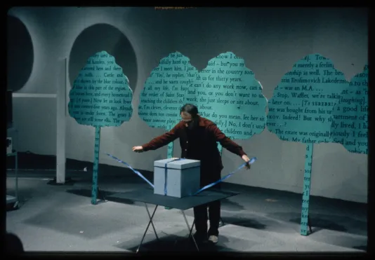 A performer stands in front of cut-out trees covered in text. The performer begins to open a blue box set on a table.